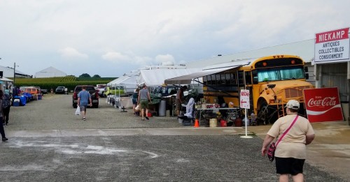 A bustling outdoor market with vendors, antique items, and a yellow school bus in the background under a cloudy sky.