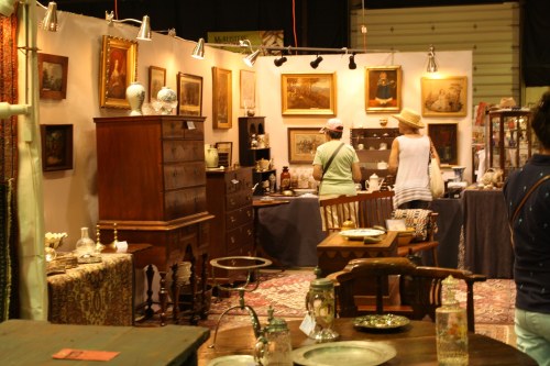 Two people browse an antique booth filled with vintage furniture, artwork, and decorative items.