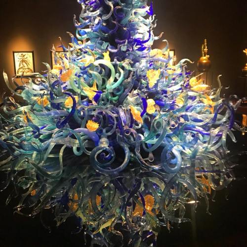 A vibrant glass chandelier with swirling blue and turquoise shapes, accented by orange and yellow elements, in a dark room.
