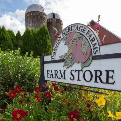 Sign for Oregon Heritage Farms Farm Store, with colorful flowers and silos in the background under a blue sky.