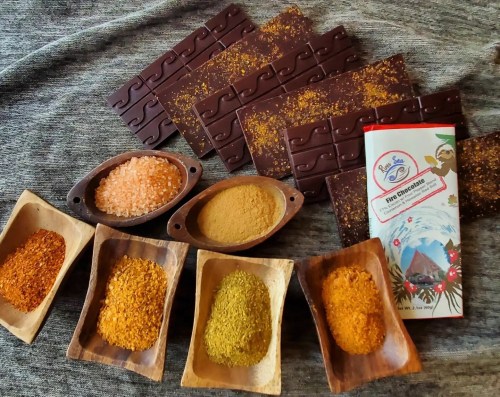 An assortment of spices in wooden bowls, pink salt, and chocolate bars arranged on a textured surface.
