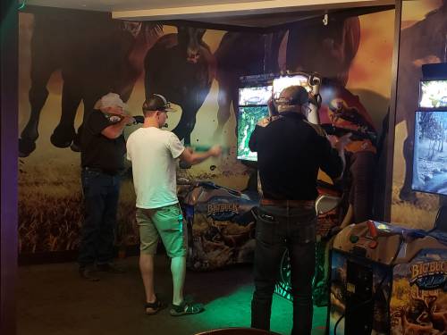Two men play arcade games in a western-themed room with horse murals on the walls.