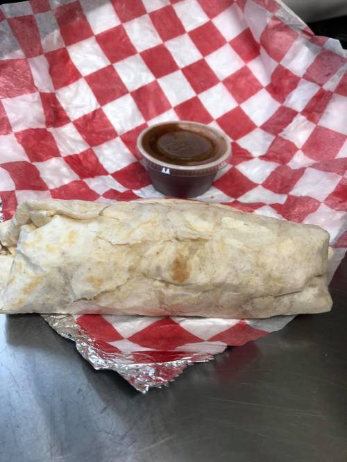A wrapped burrito on a red and white checkered paper, accompanied by a small cup of sauce.