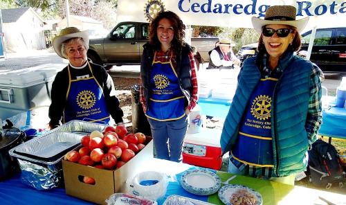Three women in blue aprons smile at a food stand with apples and baked goods, under a Rotary Club banner.