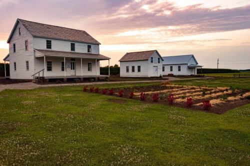Two white houses with a garden in the foreground, set against a colorful sunset sky. Green fields in the background.