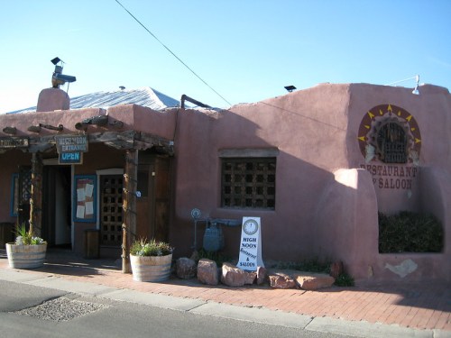 A rustic adobe building with a sign reading "Restaurant & Saloon," featuring wooden accents and potted plants outside.