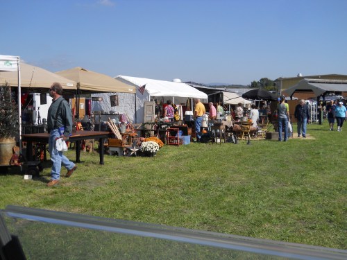 A bustling outdoor market with tents selling antiques and crafts, surrounded by people browsing and enjoying the day.