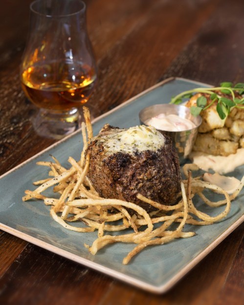 A plated dish featuring a beef filet topped with butter, served with crispy fries and a glass of whiskey in the background.