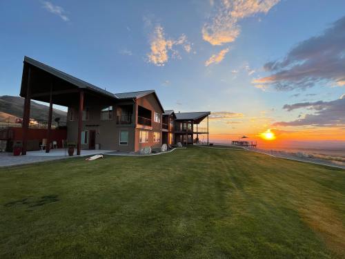 A modern house with large windows overlooks a sunset, set against a grassy landscape and distant hills.
