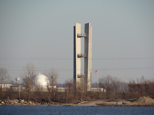 A tall, modern structure with two vertical sections near a body of water, with a dome-shaped building and trees in the background.