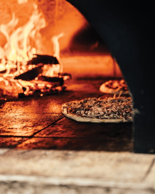 A pizza baking in a wood-fired oven, with flames visible in the background.