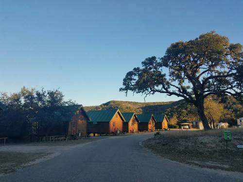 A scenic view of rustic cabins along a winding road, surrounded by trees and hills under a clear blue sky.