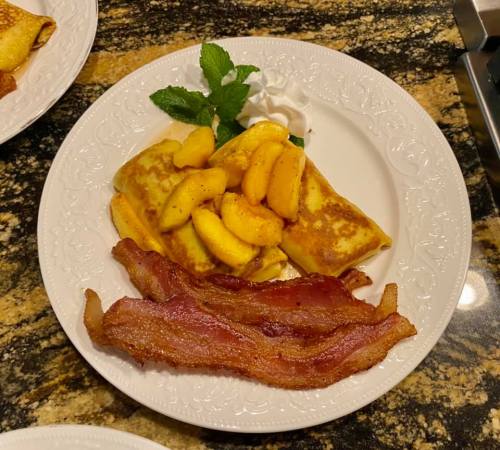 A plate with pancakes topped with sautéed apples, whipped cream, and crispy bacon, garnished with a mint leaf.