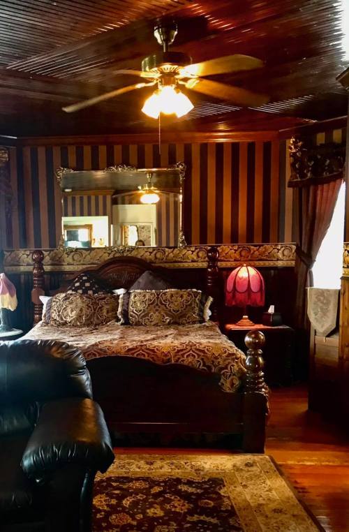 Cozy bedroom with a wooden bed, ornate decor, a ceiling fan, and a warm lamp, featuring striped walls and rich textures.