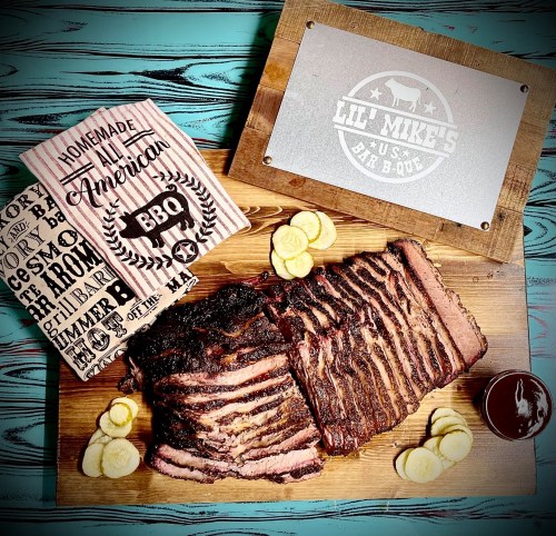 A wooden board with sliced BBQ brisket, a cookbook titled "Homemade American BBQ," and a small bowl of sauce.