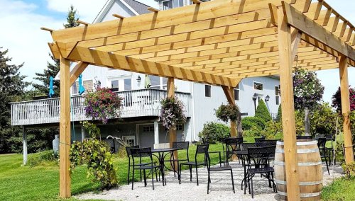 A wooden pergola with hanging plants, surrounded by tables and chairs, near a house and green lawn.
