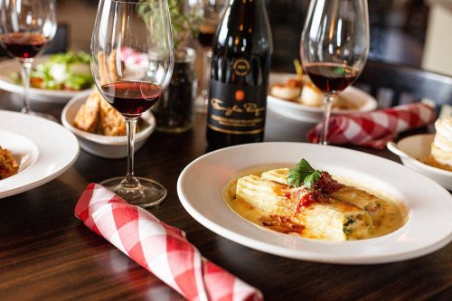 A plate of pasta with sauce and herbs, surrounded by glasses of red wine and various dishes on a dining table.
