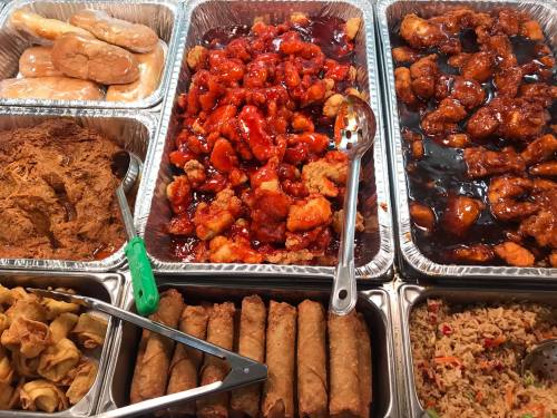 A variety of Chinese dishes including sweet and sour chicken, spring rolls, fried rice, and dumplings in metal trays.