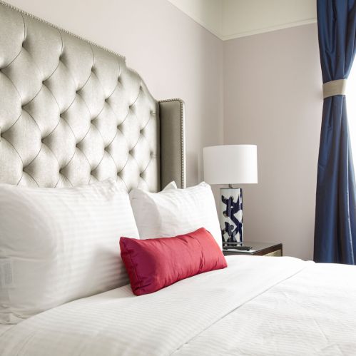 A stylish hotel bedroom featuring a tufted headboard, white bedding, a red accent pillow, and a modern lamp.