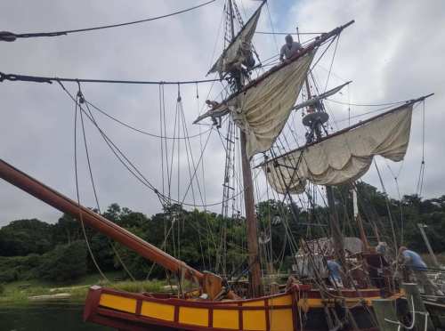 A historic sailing ship with billowing sails and crew members working on the rigging against a cloudy sky.