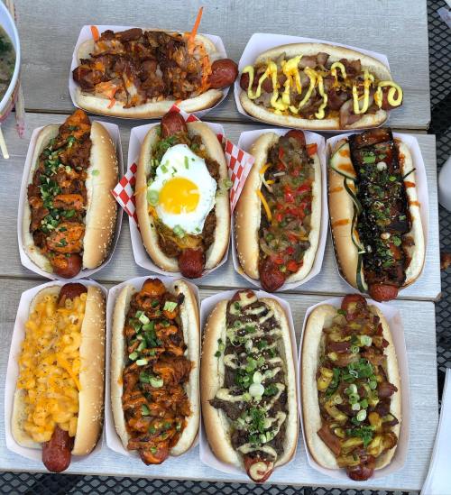 A variety of gourmet hot dogs topped with different ingredients, including meats, sauces, and vegetables, arranged on a table.