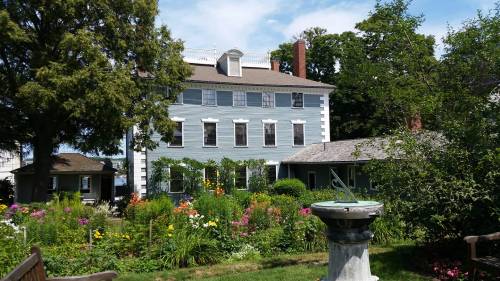A blue house surrounded by colorful flowers and greenery, with a stone sundial in the foreground.