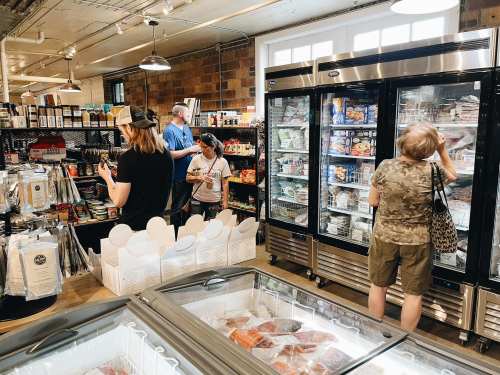 A busy grocery store interior with shoppers browsing frozen foods and shelves filled with various products.