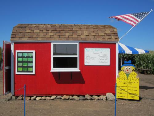 A small red building with a shingled roof, featuring a white window, a chalkboard, and a sign with a scarecrow.