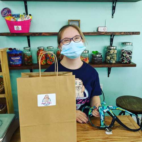 A smiling person wearing a mask stands behind a counter with a gift bag and colorful jars in the background.