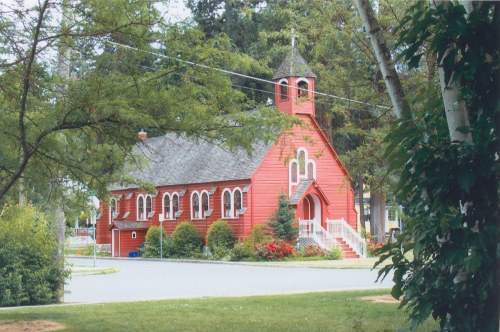 A charming red church with a steeple, surrounded by greenery and colorful flowers, set in a peaceful neighborhood.