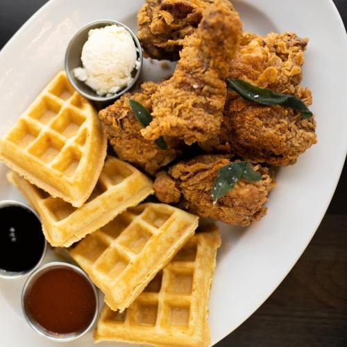 A plate of crispy fried chicken served with waffles and small bowls of syrup and sauce.