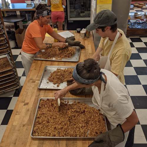 Three people in an industrial kitchen prepare granola on large trays, wearing aprons and gloves.