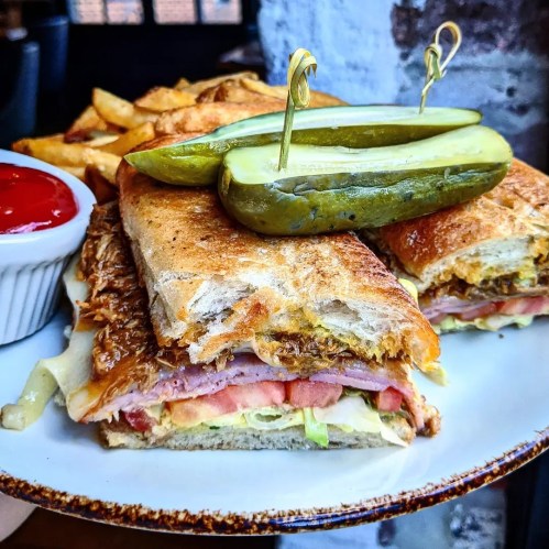 A toasted sandwich with ham, cheese, lettuce, and tomato, served with pickles and fries on the side.