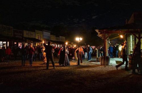 A lively night scene in a historic town, with people mingling and shops illuminated by warm lights.