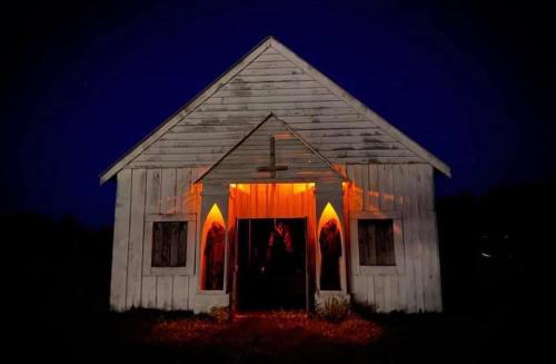 A dimly lit, old wooden church at night, with glowing orange light illuminating the entrance and windows.