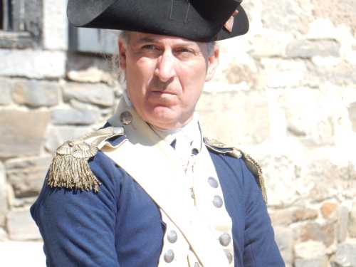 A man in a historical military uniform with a tricorn hat, looking serious against a stone wall backdrop.