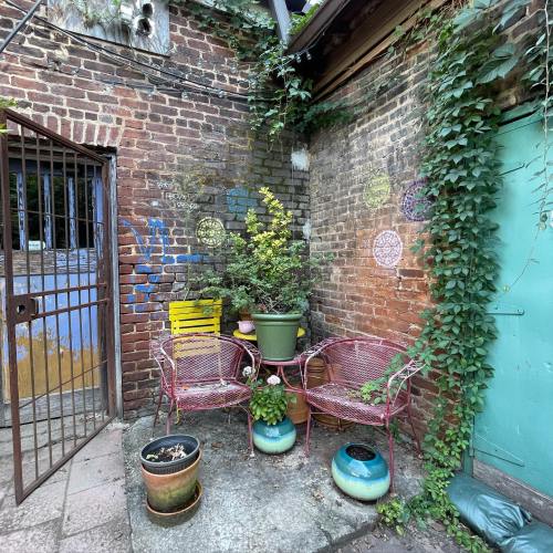 A cozy outdoor corner with two pink chairs, a potted plant, and colorful murals on a brick wall, surrounded by greenery.