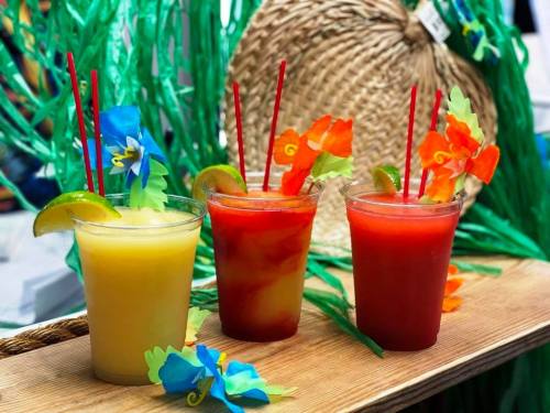 Three colorful tropical drinks with garnishes, set against a backdrop of green decorations and a straw hat.