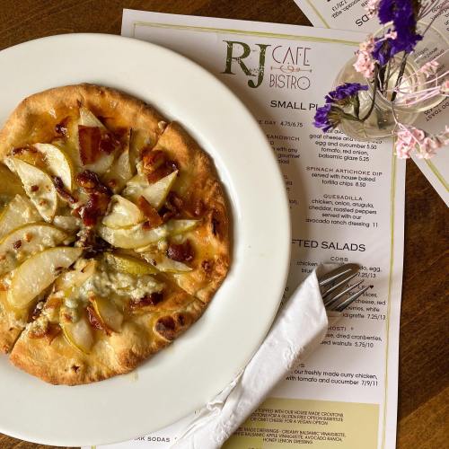 A plate with a flatbread topped with pear slices and bacon, next to a menu and a small vase of flowers.