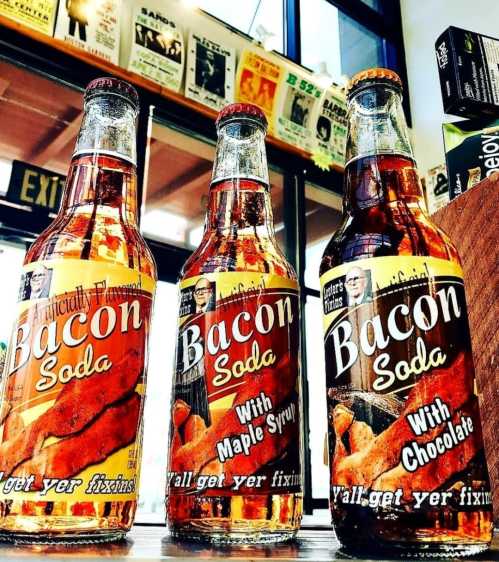 Three bottles of bacon soda in flavors: original, maple syrup, and chocolate, displayed on a counter.