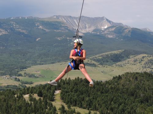 A person zip-lining over a mountainous landscape, surrounded by trees and hills under a cloudy sky.