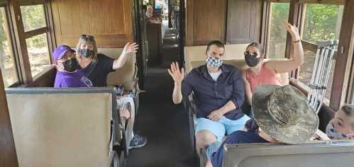 A group of people wearing masks wave from their seats on a train, smiling and enjoying their time together.