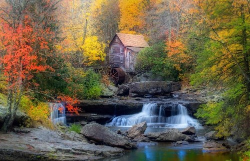 A rustic wooden mill beside a waterfall, surrounded by vibrant autumn foliage in shades of orange and yellow.