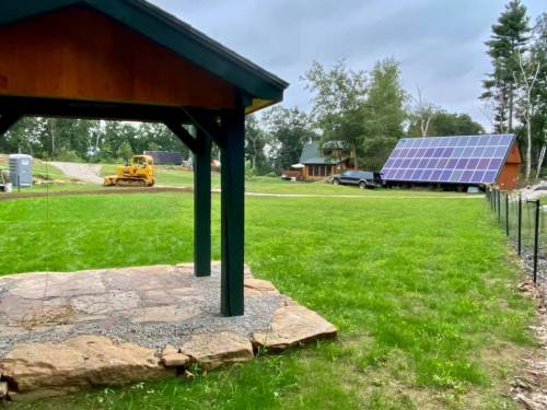 A grassy area with a stone patio, a gazebo, and a solar panel installation in the background under a cloudy sky.