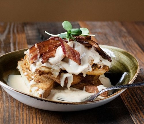A plate of fried chicken on toast, topped with crispy bacon and creamy sauce, garnished with a sprig of greens.