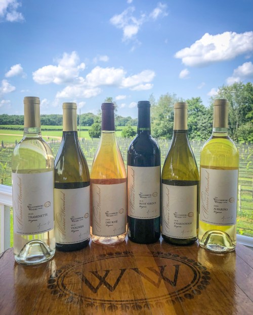 A selection of six wine bottles on a wooden table, with a vineyard and blue sky in the background.