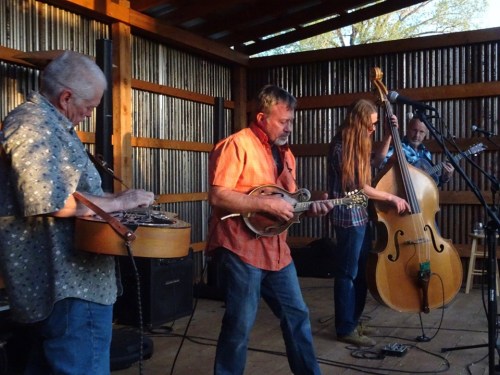 Four musicians perform on stage, playing a mandolin, banjo, and double bass in a rustic setting.