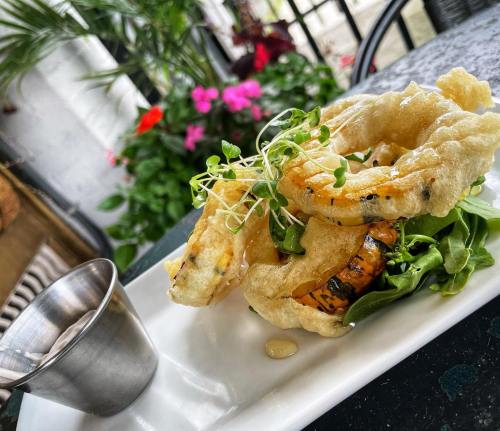 Crispy fried shrimp on a bed of greens, garnished with microgreens, served with a small metal cup of sauce.