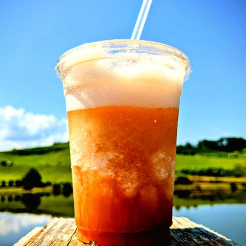 A refreshing iced drink in a clear cup, set against a sunny landscape with blue skies and green hills.