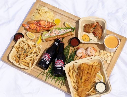 A wooden tray with seafood, fries, chips, sauces, and two bottles of root beer, all arranged on a white background.